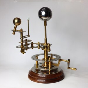 May include: A brass and silver model of the solar system with a hand crank. The model features a silver sphere representing the sun, a gold sphere representing the earth, and a smaller gold sphere representing the moon. The model is mounted on a wooden base.