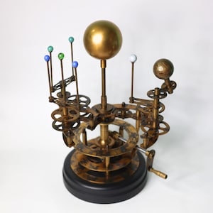 May include: A brass and gold-toned model of the solar system with a central sun and orbiting planets. The planets are represented by small colored spheres on rods. The model is mounted on a base with a crank to rotate the planets.