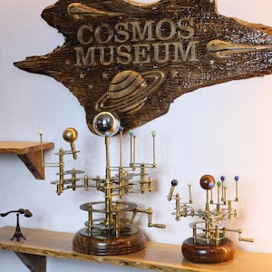 May include: Orrery models with brass gears and colorful planet spheres sit on a wooden shelf. The models are in front of a wooden sign that reads "COSMOS MUSEUM" with a planet and stars carved into it. A small Eiffel Tower model is also on the shelf.