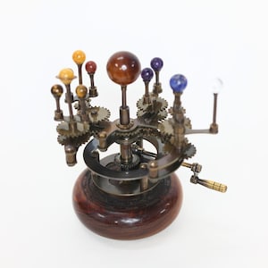 May include: A mechanical orrery model featuring a central brown sphere representing the sun, surrounded by smaller spheres of various colors and sizes on brass gear arms, all mounted on a polished dark wood circular base with a hand crank.