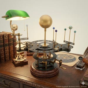 May include: A handmade mechanical orrery with a large gold sphere at the center, representing the sun. The orrery features brass rods with smaller spheres, gears, and a wooden base. A green desk lamp and antique books are in the background.