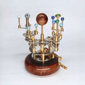 Può includere: Un modello del sistema solare in ottone e legno con una base in legno e un meccanismo rotante. I pianeti sono rappresentati da piccole sfere di vetro di vari colori, tra cui blu, verde e bianco.