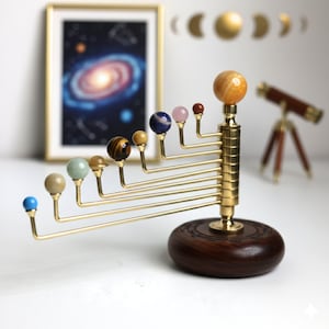 以下が含まれることがあります： 宝石の惑星と真鍮のアームを備えた装飾的な太陽系モデル。濃い色の木製のベースに取り付けられています。額入りの銀河プリントと小さな望遠鏡が背景にあります。惑星はさまざまな色とサイズです。