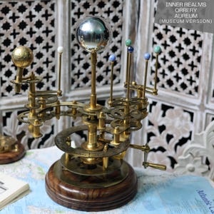 May include: A brass orrery model of the solar system with a silver-colored sun and colored planets. The orrery is on a wooden base and has a crank to rotate the planets.
