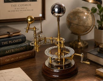 Puede incluir: Un modelo mecánico de orrery en latón que presenta una gran esfera central plateada, una esfera dorada más pequeña y un orbe blanco diminuto montados sobre una base circular con manivela. El dispositivo incluye engranajes y un dial impreso.