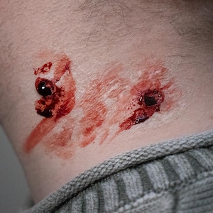 Puede incluir: Primer plano de un cuello con dos heridas simuladas, con sangre roja oscura y un efecto realista. La piel es pálida y la persona lleva un jersey gris. Ideal para Halloween o efectos especiales.