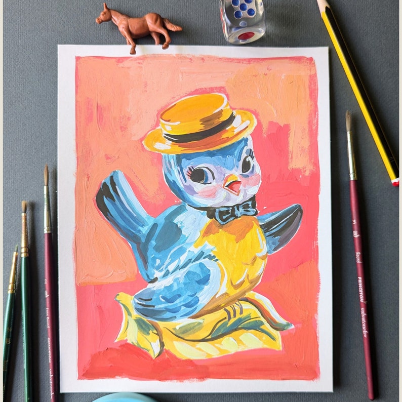 Op de afbeelding: Een blauw en geel vogelbeeldje met een gele hoed en een rode strik. De vogel zit op een geel en wit blad. De achtergrond is roze en oranje.