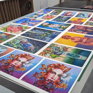 Op de afbeelding: Een verzameling kleurrijke, ingelijste prints met verschillende onderwerpen, waaronder bloemen, vogels en mensen. De prints zijn in rijen op een tafel gerangschikt, waarbij de bovenste rij het meest zichtbaar is. De prints zijn allemaal in verschillende maten en vormen, en ze zijn allemaal ingelijst in eenvoudige zwarte lijsten.