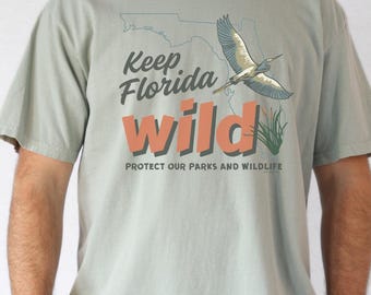 Op de afbeelding: Een saliegroen t-shirt met korte mouwen en een afbeelding van de staat Florida, een vliegende vogel en groen riet. De tekst Keep Florida wild en PROTECT OUR PARKS AND WILDLIFE staat in donkergrijs en oranje op de voorzijde gedrukt.