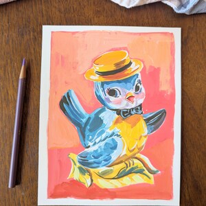 Op de afbeelding: Een handgeschilderde illustratie van een blauwe vogel die een gele hoed en een zwarte strik draagt. De vogel zit op een geel blad met een roze achtergrond.
