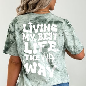 Puede incluir: Camiseta verde tie-dye con texto blanco que dice "Living my best life the NA way" con un círculo blanco que contiene las letras NA en el centro.
