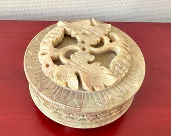 May include: A round, carved stone box with a detailed floral design. The box is made of a light brown stone with a slightly textured surface.