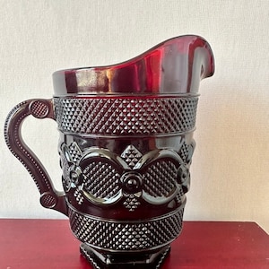 May include: A dark red glass pitcher with a diamond pattern and a decorative design around the middle. The pitcher has a handle and a hexagonal base.