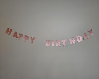 May include: A rose gold glitter "HAPPY BIRTHDAY" banner strung across a white string. The letters are individual and attached to the string. The banner is against a plain, light gray background. Perfect for a birthday party.