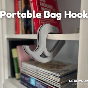 May include: A white 3D printed portable bag hook hanging on a white shelf. The hook is designed to hang on the edge of a shelf or table. The hook is made of a durable plastic and is easy to install.