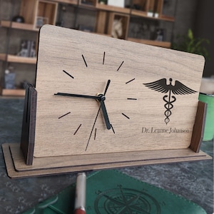 Puede incluir: Un reloj de escritorio de madera con una esfera y agujas negras. El reloj tiene un símbolo médico y el nombre "Dr. Leanne Johanson" grabado en la parte delantera.