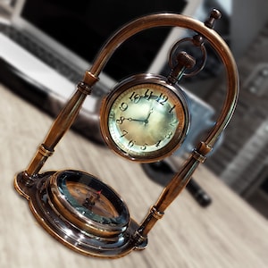 May include: A brass clock stand with a round compass face and a pocket watch with Roman numeral markings. The clock stand is arched and has a decorative design.
