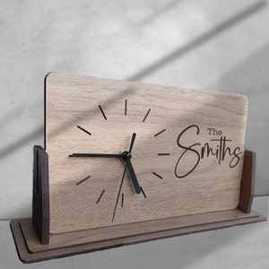 Puede incluir: Un reloj de madera con una esfera y agujas negras. El reloj tiene un soporte y el texto "The Smiths" está grabado en la esfera.