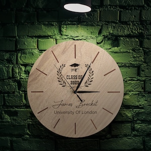 Peut inclure: Une horloge en bois avec un design de chapeau de graduation et une couronne de laurier. L'horloge indique "Classe de 2023" et porte le nom "James Brocket" et "Université de Londres" inscrit en dessous.