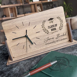 May include: Wooden desk clock with a graduation cap and laurel wreath design. The clock reads "Class of 2023" and is personalized with the name "James Brocket" and "University of London".