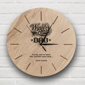 Peut inclure: Une horloge en bois avec un fond brun et des aiguilles noires. Le cadran de l'horloge affiche le texte "World's Best Dad" avec des étoiles de chaque côté du mot "Dad". L'horloge affiche également le texte "Lucky me to have the world's best dad... Love Lucie".