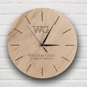 以下が含まれることがあります： 明るい茶色の木製丸時計。文字盤にはイニシャル「WG」と「WILLIAM GRAY Creative Director」の名前が黒色で表示されています。茶色の線が時間を表し、時計の針は黒色です。