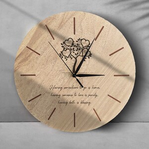 Könnte beinhalten: Eine Wanduhr aus Holz mit einem Baum- und Herzdesign. Die Uhr hat schwarze Zeiger und braune römische Ziffern. Der Spruch "Einen Ort zum Hingehen zu haben, ist Zuhause, jemanden zu lieben, ist Familie, beides zu haben, ist ein Segen." ist auf dem Zifferblatt der Uhr gedruckt.