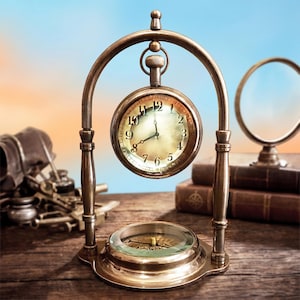 Puede incluir: Un reloj de escritorio vintage de latón con base de brújula y esfera de reloj de bolsillo. El reloj tiene un soporte curvo y un pequeño lazo en la parte superior. La esfera tiene números romanos negros y agujas doradas. El reloj está sobre una superficie de madera.