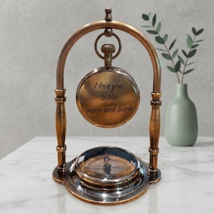 Puede incluir: Una brújula de latón de estilo vintage con un arco decorativo y detalles de madera. La brújula tiene una esfera redonda con una aguja y una cubierta de vidrio. El texto "I love you to the moon and back" está inscrito.