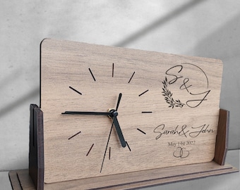 May include: A wooden desk clock with a black clock face and hands. The clock has a personalized inscription that reads "Sarah & John May 1st 2022" with a decorative wreath and initials "S&J".