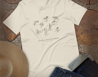 Puede incluir: Una camiseta de manga corta color crema con un gráfico de líneas negras de varias ilustraciones botánicas. Debajo de las plantas, el texto NATIVE WILDFLOWERS OF MICHIGAN aparece en una fuente sans-serif clara centrada en el pecho.