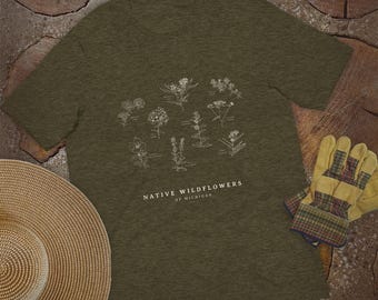Puede incluir: Una camiseta de manga corta color verde oliva con una ilustración botánica blanca de varias plantas y el texto "NATIVE WILDFLOWERS OF MICHIGAN" impreso en el centro. La camiseta tiene cuello redondo y una textura de tela jaspeada.