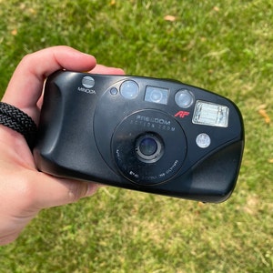 Könnte beinhalten: Eine schwarze Minolta Freedom Action Zoom AF Kamera mit einem 38mm Objektiv. Die Kamera verfügt über einen eingebauten Blitz und ein Zoomobjektiv.