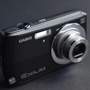 Pode incluir: Uma câmera digital Casio Exilim preta com uma lente prateada e um botão prateado na parte superior. A câmera possui um flash embutido e um zoom.
