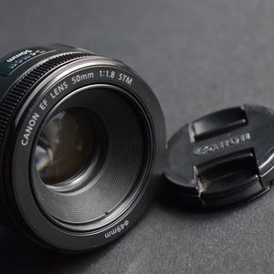 Può includere: Un obiettivo Canon EF 50mm f/1.8 STM nero con il tappo dell'obiettivo rimosso. L'obiettivo è un obiettivo standard per le fotocamere reflex digitali Canon.