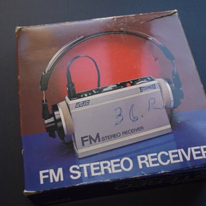 Könnte beinhalten: Vintage FM Stereo Receiver in der Originalverpackung. Die Box ist blau und rot mit dem Produktbild. Der Empfänger ist silberfarben mit schwarzen Akzenten und hat einen Kopfhörer. Der Text "FM STEREO RECEIVER" ist auf der Box aufgedruckt.