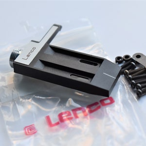 May include: A black Lenco turntable headshell with a silver trim and a white line. The headshell is packaged in a clear plastic bag with a red Lenco logo. The package also includes black screws and a black metal bracket.