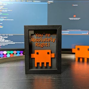 May include: A framed art piece with the text "You're Absolutely Right!" in orange. The frame is black, and the artwork features an orange pixelated creature. Additional orange pixelated creatures are on the desk.