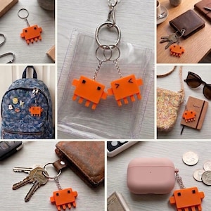 A collage of nine images shows the orange, pixelated crab-like keychain attached to various items like backpacks, keys, lanyards, and a wallet.