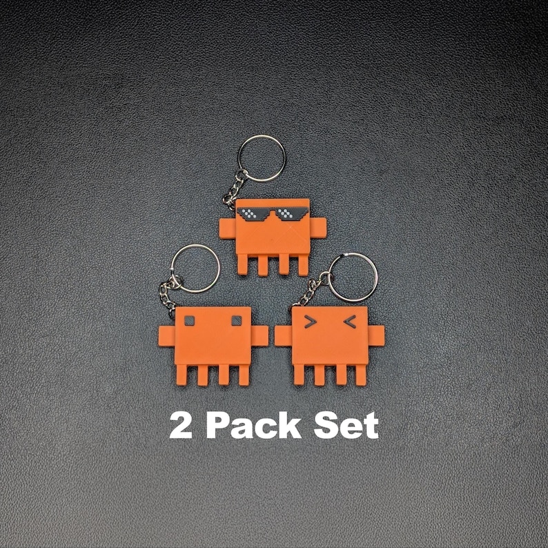 Three orange, pixelated crab-like keychains, two with V-shaped eyes and one with sunglasses, are arranged on a dark gray surface with "2 Pack Set" text below them.