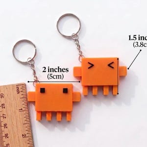 Two orange, pixelated crab-like keychains are shown next to a ruler, indicating they are 2 inches (5cm) wide and 1.5 inches (3.8cm) tall.