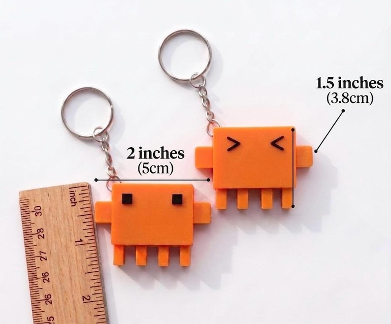 Two orange, pixelated crab-like keychains are shown next to a ruler, indicating they are 2 inches (5cm) wide and 1.5 inches (3.8cm) tall.