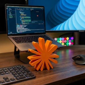 May include: An orange, star-shaped object rests on a wooden desk. A laptop displaying code is in the background, alongside a keyboard and mouse. The object's design features multiple radiating blades.