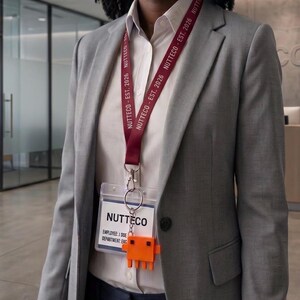 May include: A maroon lanyard with the white text NUTTECO - EST. 2026 attached to a clear plastic ID badge holder. The badge displays the text NUTTECO, EMPLOYEE: J. DOE, and DEPARTMENT: ENG. An orange pixelated character charm hangs from the metal ring.