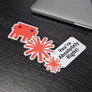 Może przedstawiać: Zestaw czerwonych i białych naklejek na ciemnej powierzchni. Naklejki zawierają postać z gry retro, dwa wzory gwiazd i naklejkę z napisem "You're Absolutely Right!"