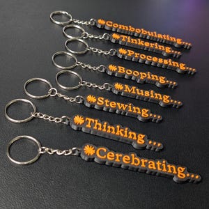 May include: A selection of keychains with black bases and orange text. Each keychain features a different word, such as "Combobulating," "Tinkering," and "Processing." The keychains are attached to silver key rings and chains.