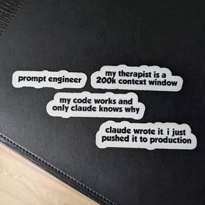 Op de afbeelding: Vier witte rechthoekige stickers met zwarte tekst. De stickers bevatten de teksten: prompt engineer, my therapist is a 200k context window, my code works and only claude knows why, en claude wrote it. i just pushed it to production.