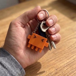 A hand holds a set of keys with an orange, pixelated crab-like keychain attached, showing its V-shaped eyes and mouth.