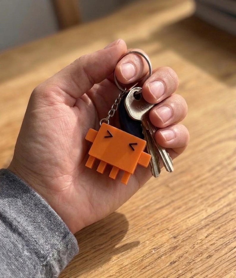 A hand holds a set of keys with an orange, pixelated crab-like keychain attached, showing its V-shaped eyes and mouth.