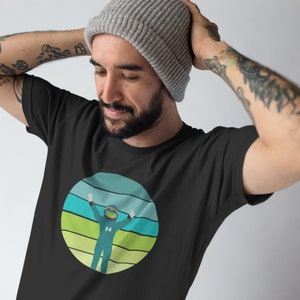 Puede incluir: Camiseta negra con un gráfico retro de un piloto de carreras con el número 14 en su traje, levantando los brazos en señal de victoria. El gráfico está sobre un fondo de rayas azules, verdes y amarillas.